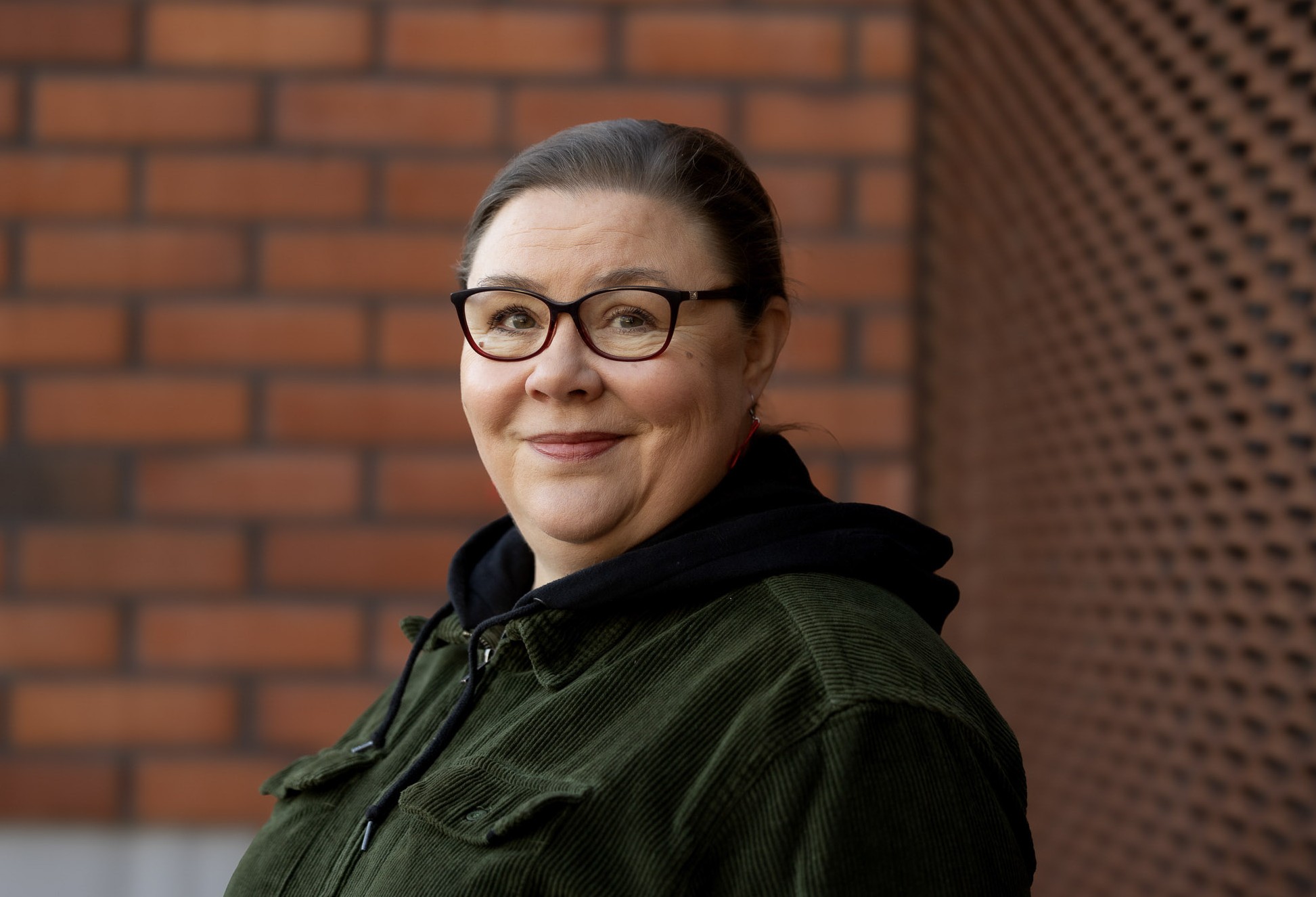 Katri Kujanpää 2026. Kuvassa hänellä on vihreä vakosamettitakki, jossa on huppu. Hän seisoo keskiruskean tiiliseinän edessä Kankaanpään liikuntakeskuksen pihassa.